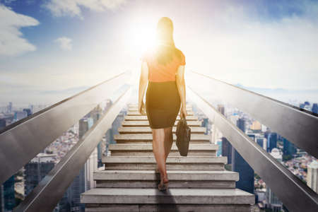 Rear view of female manager carrying a suitcase while climbing stairs toward bright sunlight with modern city backgroundの写真素材