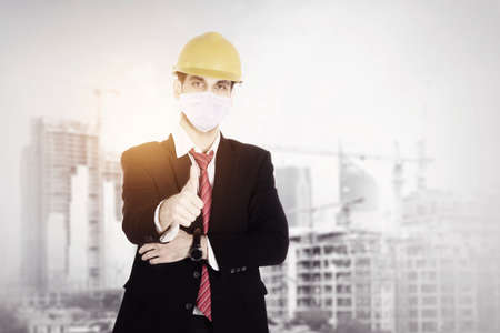 Double exposure of male contractor wearing face mask while showing thumbs up and standing with...