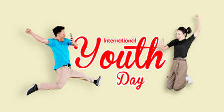 Two young people using smartphone while jumping with International youth day text in the studio with cream backgroundの写真素材