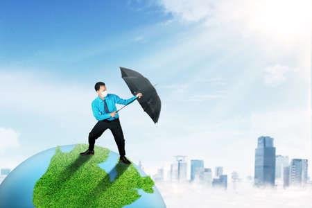 Young businessman with an umbrella protect planet Earth from ozone depletion effects while wearing face maskの写真素材