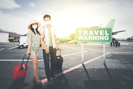 Young couple wearing face mask while standing with travel warning text on the signpost at airport...