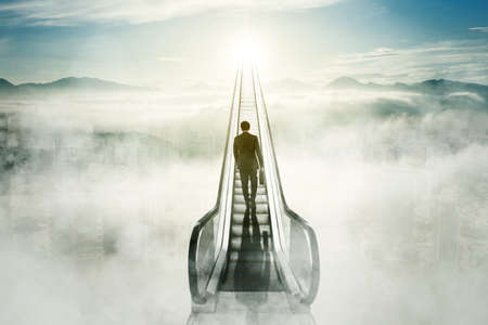 Rear view of male manager carrying a suitcase while walking upward on the escalator with misty city...