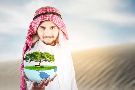 Arabian man holding an earth planet and green tree while standing in the desertの写真素材