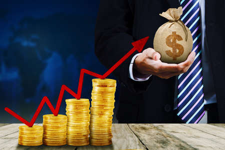 Businessman hands holding money bag while standing with upward arrow and coins shaping growth graph in world map backgroundの写真素材