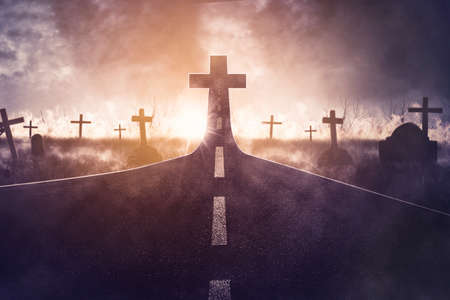 Graveyard with empty road with cross sign on end the road at sunset time. Concept of Halloween...