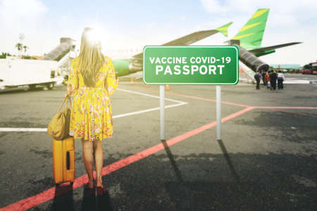 Blonde woman carrying a luggage and handbag while standing with vaccine covid-19 passport text in the airportの写真素材