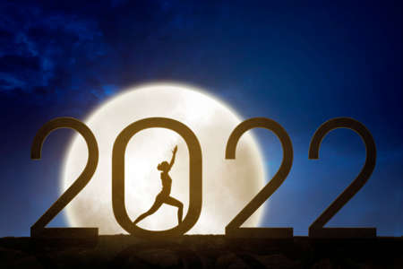 Silhouette of young woman practicing yoga while standing with 2022 numbers and full moon background at night timeの写真素材
