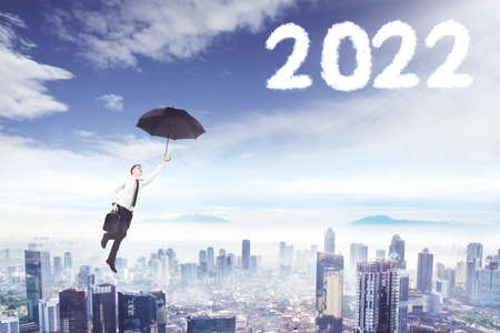 Young businessman using an umbrella while flying toward clouds shaped numbers 2022 in the blue skyの写真素材