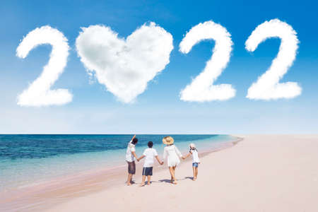 Back view of young family holding hands together on the beach while looking at clouds shaped 2022...