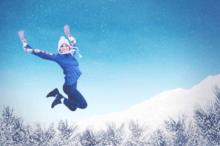 Excited young woman jumping at the park under snowfall while wearing winter clothes at Christmas...