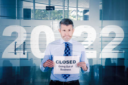 Sad businessman holding text of closed going out of business on the paper while standing with 2022 numbers on the office windowの写真素材