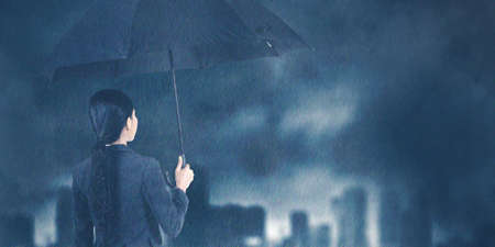 Back view of businesswoman holding an umbrella while standing under raining with cloudy sky backgroundの写真素材