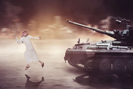 Middle Eastern man looks scared while running away from a military tank with ruined city backgroundの写真素材