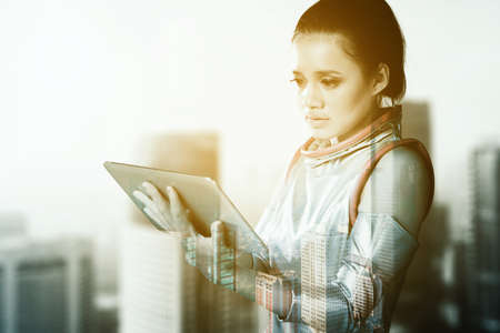 Double exposure of futuristic woman using a digital tablet while standing with cityscape background