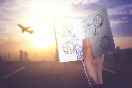 Close up of young man hand holding a stamped passport while standing with silhouette of airplane at sunset timeの写真素材