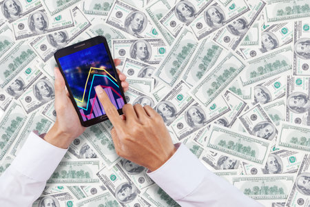 Close up of businessman hands touching finance graph on a cellphone with stacks of moneyの写真素材