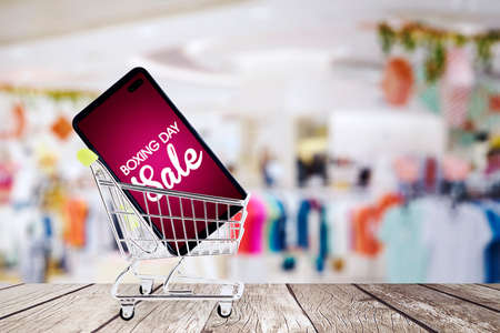Mini shopping cart carrying a cellphone with boxing day sale text on the wooden table. Shot with clothing store backgroundの写真素材