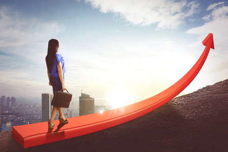 Young businesswoman carrying a suitcase while walking above upward arrow on the hill with cityscape backgroundの写真素材