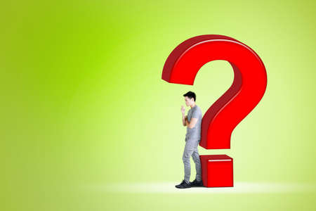 Confused young man thinking something while standing with question mark in the studio with green...