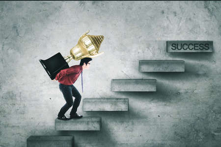 Image of businessman carrying a trophy while climbing stairs toward success wordの写真素材