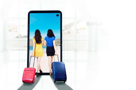 Rear view of two young women carrying luggage while walking toward tropical beach on the cellphone screenの写真素材