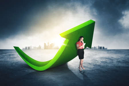 Pensive businesswoman holding a mobile phone while standing near a green upward arrow with skyscraper backgroundの写真素材