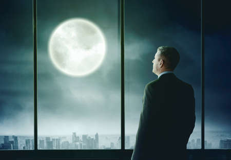 Rear view of businessman looking at the moon at night time while standing in the officeの写真素材