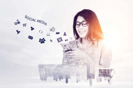 Double exposure of happy businesswoman using a cellphone with social media icon while sitting with cityscape backgroundの写真素材