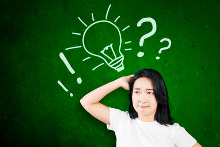 Confused woman scratching her head while looking at drawn bright light bulb on the chalkboardの写真素材