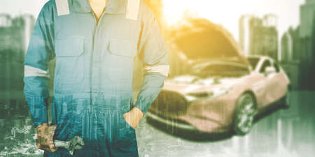 Double exposure of male mechanic holding a wrench while standing with broken car and cityscape backgroundの写真素材