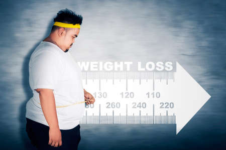 Side view of obesity man measuring his belly with measuring tape while standing with weight loss text backgroundの写真素材