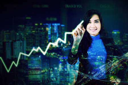 Double exposure of businesswoman drawing upward arrow while standing with night city background
