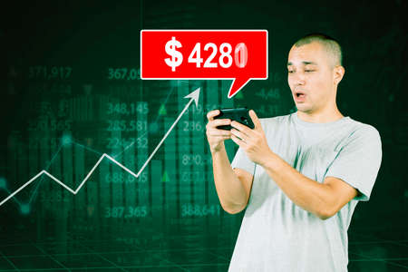 Surprised young man looking at increasing stock market price on the cellphone while standing with...