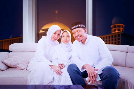Happy Muslim family smiling at the camera while sitting together on the sofa during Eid Mubarak with mosque backgroundの写真素材