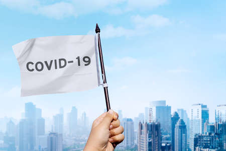 Close up of unknown hand waving a white flag with Covid-19 word while standing with cityscape backgroundの写真素材