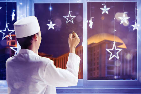 Young man praying to the Allah and holding beads while standing near the window with mosque view