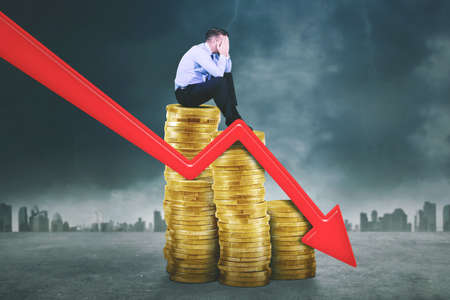 Businessman looks sad while sitting on piles of coins near declining arrow with cloudy sky...
