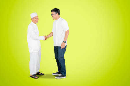 Elderly man handshaking with his son at Eid Mubarak celebration while standing in the studio with...