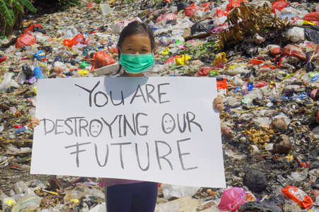 JAKARTA - Indonesia. April 26, 2022: Little girl wearing a mask and showing a banner with text of you are destroying our future while standing in the landfillの写真素材