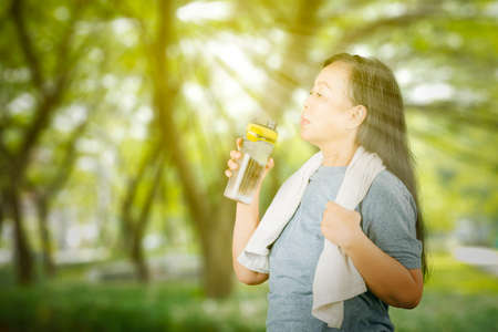 Side view of senior woman drinking a bottle of water after exercise at the park with sunlight...