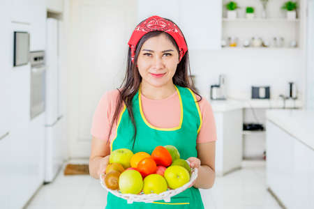 Pretty housewife holding a basket of fresh fruit in the kitchen while looking and smiling at the camera. Shot at homeの写真素材