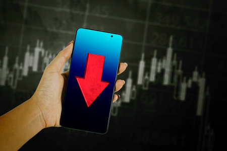 Young man hand showing a falling arrow on the cellphone with declining candlestick chart backgroundの写真素材
