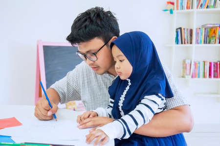 Young man teaching his daughter to write on a book while learning together at homeの写真素材