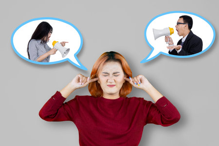Angry irritated asian woman covers her ears with hands while standing isolated over gray background
