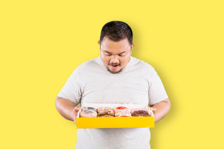 Overweight fat Asian man holding a box with colorful glazed doughnuts isolated over yellow backgroundの写真素材