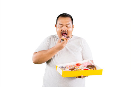 Overweight fat Asian man eating a doughnut while the hand holding a box with colorful glazed doughnuts isolated over whiteの写真素材
