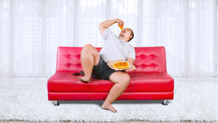 Overweight Asian guy eating a box of pizza while sitting on a couch isolated at homeの写真素材