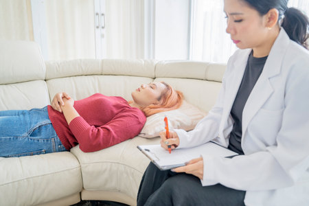 Psychotherapist listening attentively to Asian young woman lying down on a couch and writes in clipboardの写真素材