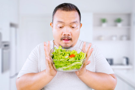 Fat Asian guy holds fresh greens salad in bowl in the kitchenの写真素材
