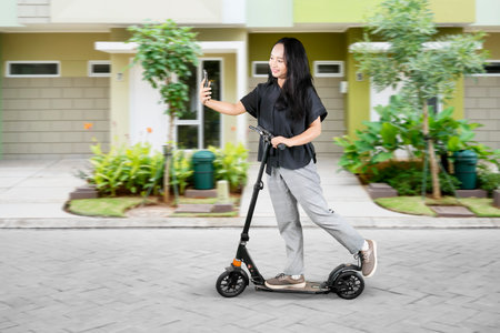 Full length profile shot of a female student with a mobile phone taking a selfie while riding an electric scooterの写真素材
